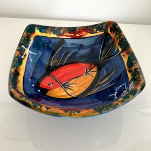 Peut inclure: Un bol carré en céramique présentant un motif peint à la main d'un poisson stylisé au corps rouge et orange avec des nageoires noires. L'intérieur a un fond bleu profond, tandis que le large bord évasé est orné de tons tachetés vert, jaune et orange.