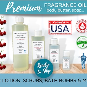 May include: Four bottles of fragrance oil labeled "The Soap Guy Premium Fragrance Oil" in various scents: Patchouli, Oatmeal, Milk & Honey, Cherry Almond, and Black Honey. The bottles are on a wooden surface with a background of red cherries and a sign that reads "Premium Fragrance Oil body butter, soap... Made in USA Alcohol 0% Free Non-Toxic Phthalate Free".
