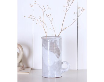 Può includere: Un vaso cilindrico in ceramica fatto a mano con manico, con smalto bianco e grigio. Il vaso contiene steli secchi e sottili con piccole capsule di semi. Il vaso è alto circa 20 cm e largo 10 cm.