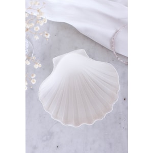Può includere: Un piatto in ceramica bianca a forma di conchiglia di mare con una texture a coste. Il piatto è perfetto per contenere piccoli oggetti come gioielli o ciondoli.