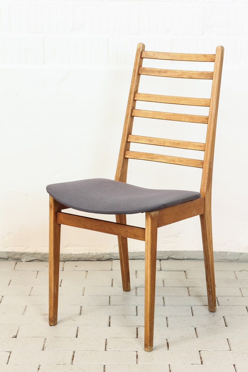 K&ouml;nnte beinhalten: Ein Holzstuhl mit einer Lattenlehne und einem grauen Stoffbezug. Der Stuhl ist aus hellem Holz gefertigt und hat ein schlichtes, klassisches Design.