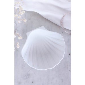 Könnte beinhalten: Eine weiße, muschelförmige Schale mit strukturierter Oberfläche. Die Schale steht auf einer hellgrauen Oberfläche, mit einem weißen Tuch und einem Perlenarmband im Hintergrund. Zarte weiße Blüten sind ebenfalls zu sehen.