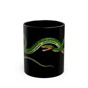 Puede incluir: Una taza de café negra con una serpiente verde y amarilla envuelta alrededor. La serpiente tiene una boca amarilla brillante con colmillos rojos.