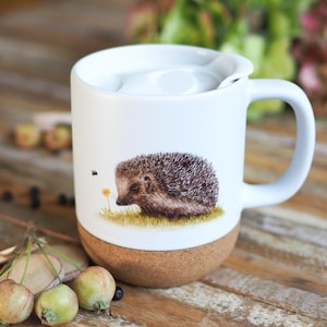 Könnte beinhalten: Weißer Keramikbecher mit Korkboden und Deckel. Der Becher zeigt eine detaillierte Illustration eines Igels auf einer Grasfläche mit einer kleinen gelben Blume. Der Becher hat einen weißen Griff.