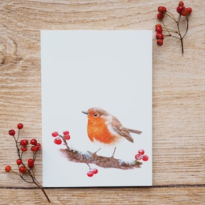 Könnte beinhalten: Aquarellmalerei eines Rotkehlchens auf einem schneebedeckten Ast mit roten Beeren. Der Vogel hat eine leuchtend orangefarbene Brust und einen braunen Rücken. Das Kunstwerk befindet sich auf einer weißen Karte, mit Zweigen roter Beeren auf Holzgrund.