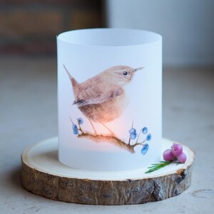Könnte beinhalten: Zylindrischer Kerzenhalter aus mattiertem Glas mit einer Aquarellillustration eines kleinen braunen Vogels, der auf einem Ast mit blauen Beeren sitzt. Der Kerzenhalter steht auf einer runden Basis aus naturbelassenem Holz.