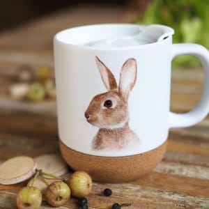 Könnte beinhalten: Weiße Keramik-Tasse mit einer detaillierten Illustration eines braunen Hasen. Die Tasse hat einen Korkboden und einen weißen Deckel. Der Griff ist weiß und die Tasse steht auf einer Holzoberfläche mit kleinen dekorativen Elementen.