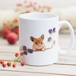 Könnte beinhalten: Weiße Keramiktasse mit einer braunen Maus, die hinter einem Zweig mit violetten Beeren hervorlugt. Die Maus hat eine rosa Nase und Schnurrhaare.