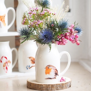 Könnte beinhalten: Eine weiße Keramikkrug mit Henkel, verziert mit einer Rotkehlchen-Illustration, enthält einen Blumenstrauß aus blauen Disteln und rosa Beeren. Der Krug steht auf einer Holzscheibe. Im Hintergrund sind weitere Krüge mit Tier- und Festtagsmotiven zu sehen.