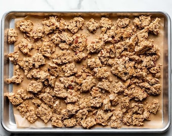 May include: A rectangular metal baking sheet lined with brown parchment paper, filled with clusters of golden-brown toasted granola. The granola consists of rolled oats and visible nut pieces, spread across the surface in uneven, crunchy chunks.