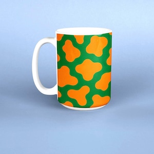 May include: White ceramic mug with a green background and orange abstract shapes. The mug has a white handle and a solid white interior. The mug is set against a light blue background.