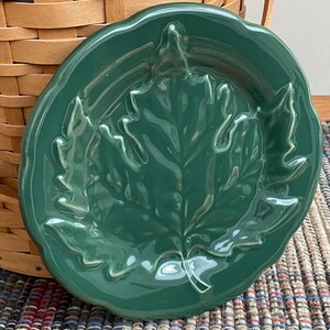 May include: A green ceramic leaf-shaped dish. The dish has a raised leaf design in the center and a scalloped edge. The dish is sitting on a woven placemat and in front of a wicker basket.