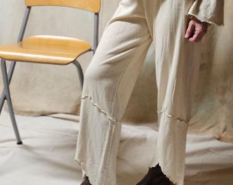 May include: Cream-colored knitwear set, including a long-sleeved top and flared pants. The pants have a unique scalloped edge design. The model is wearing brown leather boots. A vintage wooden chair with a metal frame is in the background.