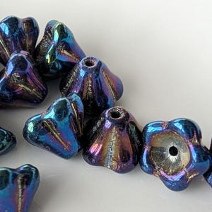 May include: A collection of iridescent blue and purple flower-shaped beads. Each bead has a hole for stringing and a textured, multi-faceted surface. The beads are scattered on a white surface.