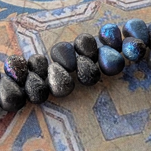 May include: A strand of teardrop-shaped beads with a dark, metallic, and iridescent finish. The beads are strung on a clear filament, displaying shades of charcoal, deep blue, and purple hues across their textured, reflective surfaces.