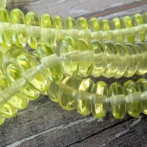 May include: Close-up of several strands of translucent, light green beads. The beads have a spiral shape and are strung together. The background is a weathered, gray wooden surface. The beads are a pale, yellowish-green color.