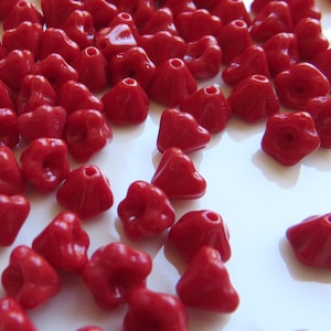 May include: A collection of small, vibrant red beads with a unique, flower-like shape. These beads are scattered across a white surface, showcasing their glossy finish and intricate design. Ideal for jewelry making or craft projects.