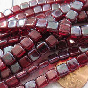 May include: Strands of square, dark red beads with iridescent highlights. The beads are arranged in rows, with a penny for size comparison. The beads have a glossy finish and are likely for jewelry making.