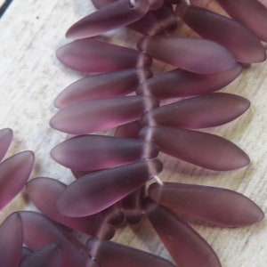 May include: A strand of purple glass beads in a leaf shape. The beads are strung on a white thread and are arranged in a staggered pattern.