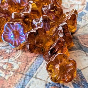 May include: A collection of amber-colored glass flower beads. The beads have a translucent quality, with some displaying iridescent hues of purple and blue. The floral design is detailed, and the beads are scattered on a patterned surface.