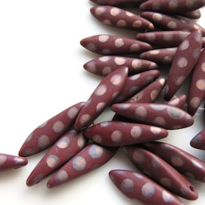 May include: A group of maroon-colored ceramic beads with white polka dots. The beads are shaped like elongated ovals and have a glossy finish.