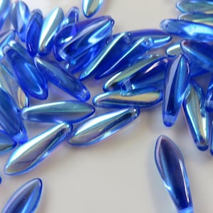 May include: A pile of iridescent blue glass beads in a teardrop shape. The beads have a pearlescent finish and are scattered on a white surface.