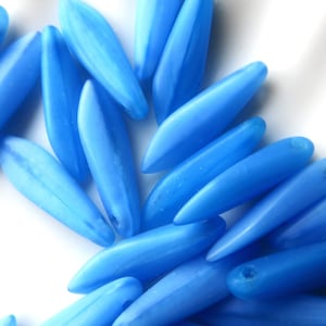 May include: A collection of light blue glass beads in a teardrop shape. The beads are scattered on a white surface.