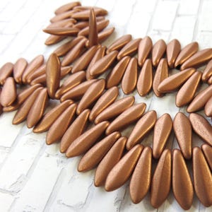 May include: A close-up of a pile of copper-colored glass beads. The beads are shaped like long, narrow teardrops. They are arranged in a circular pattern.