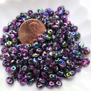 May include: A pile of iridescent beads in shades of purple, blue, and green. A copper penny rests among the beads. The beads are small and have a glossy finish, perfect for jewelry making or crafts.