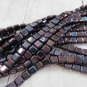 May include: Strands of square, dark purple beads with a metallic sheen. The beads are strung together, creating a textured and reflective surface. The beads are arranged in a diagonal pattern.