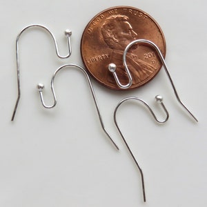 May include: Three silver earring hooks with small round balls at the end. The hooks are shown next to a US penny for size comparison.