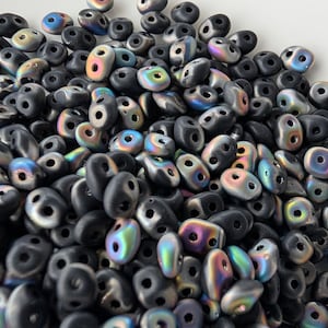 May include: A pile of black, two-hole beads with an iridescent finish. The beads have a smooth, rounded shape and a matte finish. The iridescent beads reflect colors like blue, green, and purple. These beads are suitable for jewelry making and crafts.