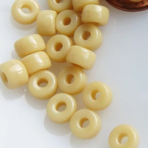 May include: A collection of light yellow, donut-shaped beads. The beads have a smooth, glossy finish and a hole in the center. They are scattered on a white surface, suggesting they are for crafting or jewelry making.