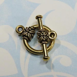 May include: A gold-colored toggle clasp with floral details. The clasp features a circular ring with a decorative floral element and a bar that inserts into the ring. The background is a light blue and white pattern.