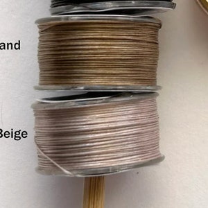 May include: Two spools of thread, one beige and one sand colored, are stacked on top of each other. The spools are on a white surface.