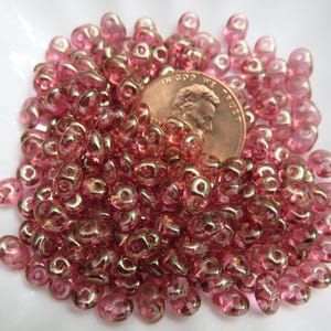 May include: A collection of pink glass beads with a metallic sheen, scattered around a U.S. penny. The beads have a unique, irregular shape and a translucent quality, creating a sparkling effect. The penny provides a size reference.
