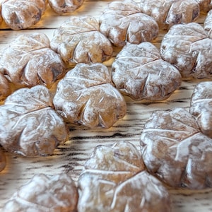 May include: Close-up of several leaf-shaped candies, individually wrapped in clear cellophane. The candies are a light brown color with a dusting of white powder, arranged on a light-colored wooden surface.