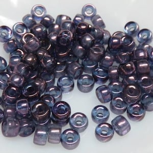 May include: A pile of small, cylindrical beads in shades of purple and blue. The beads have a glossy, iridescent finish, reflecting light. These beads are suitable for jewelry making and crafting projects.