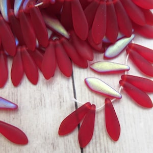 May include: Close-up of red teardrop-shaped beads with a matte finish, strung together on white thread. Some beads have an iridescent, pearlescent sheen. These beads are ideal for jewelry making and crafting projects.