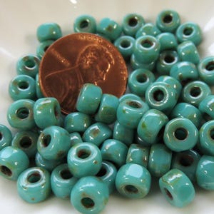 May include: A collection of turquoise blue glass beads with a slightly distressed finish. The beads are round and have a hole in the center. They are arranged on a white surface.