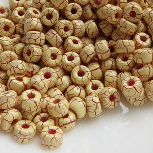 May include: A pile of cream-colored beads with a cracked, aged appearance. The beads have a reddish-brown crackle pattern and a central hole. These beads are suitable for jewelry making and crafts.