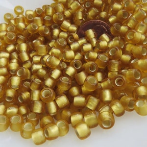 May include: A pile of translucent, golden-colored glass beads. The beads are cylindrical with a hole running through the center. A U.S. penny is visible among the beads, providing a size reference. The beads have a slightly rounded shape.
