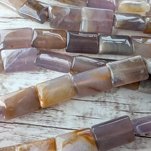 May include: Close-up of a strand of rectangular gemstone beads in shades of purple, brown, and cream. The beads are polished and translucent, with variations in color and pattern. The beads are strung together, ready for jewelry making.
