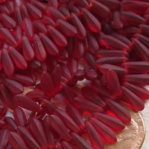 May include: Close-up of a pile of translucent, teardrop-shaped beads in a deep red hue. The beads are clustered together, with a penny visible for size comparison. The beads have a smooth, glossy finish, suggesting they are made of glass or a similar material. Ideal for jewelry making.