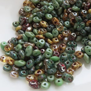 May include: A collection of colorful beads in shades of green, brown, and iridescent hues. The beads have a variety of shapes, including oval and double-holed designs. The beads are scattered, creating a textured and visually appealing display, perfect for jewelry making.