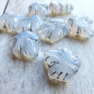 May include: A group of opaque white glass beads in the shape of leaves with a silver-colored design. The beads are scattered on a white wooden surface.