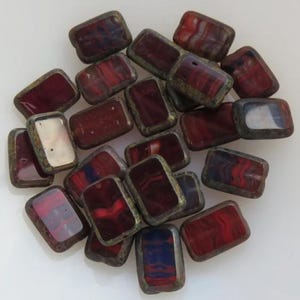 May include: A collection of rectangular beads with a dark red and blue swirled pattern. The beads have a metallic bronze-colored border. The beads are a mix of opaque and translucent. The beads are scattered on a white surface.