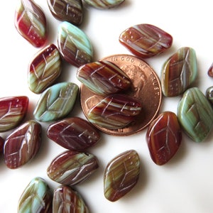 May include: A collection of glass beads in shades of brown, red, and blue, with a leaf design. The beads are scattered on a white surface, with a penny for scale.