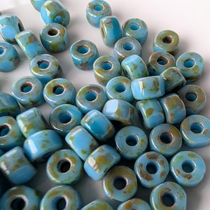 May include: A collection of small, round, ceramic beads featuring a glossy light blue glaze with irregular brown and tan mottled spots. Each bead has a central hole and a smooth, rounded shape, scattered across a plain white surface.