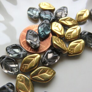 May include: A collection of glass and metal leaf-shaped beads in shades of gold, gray, and teal. The beads are scattered on a white surface.